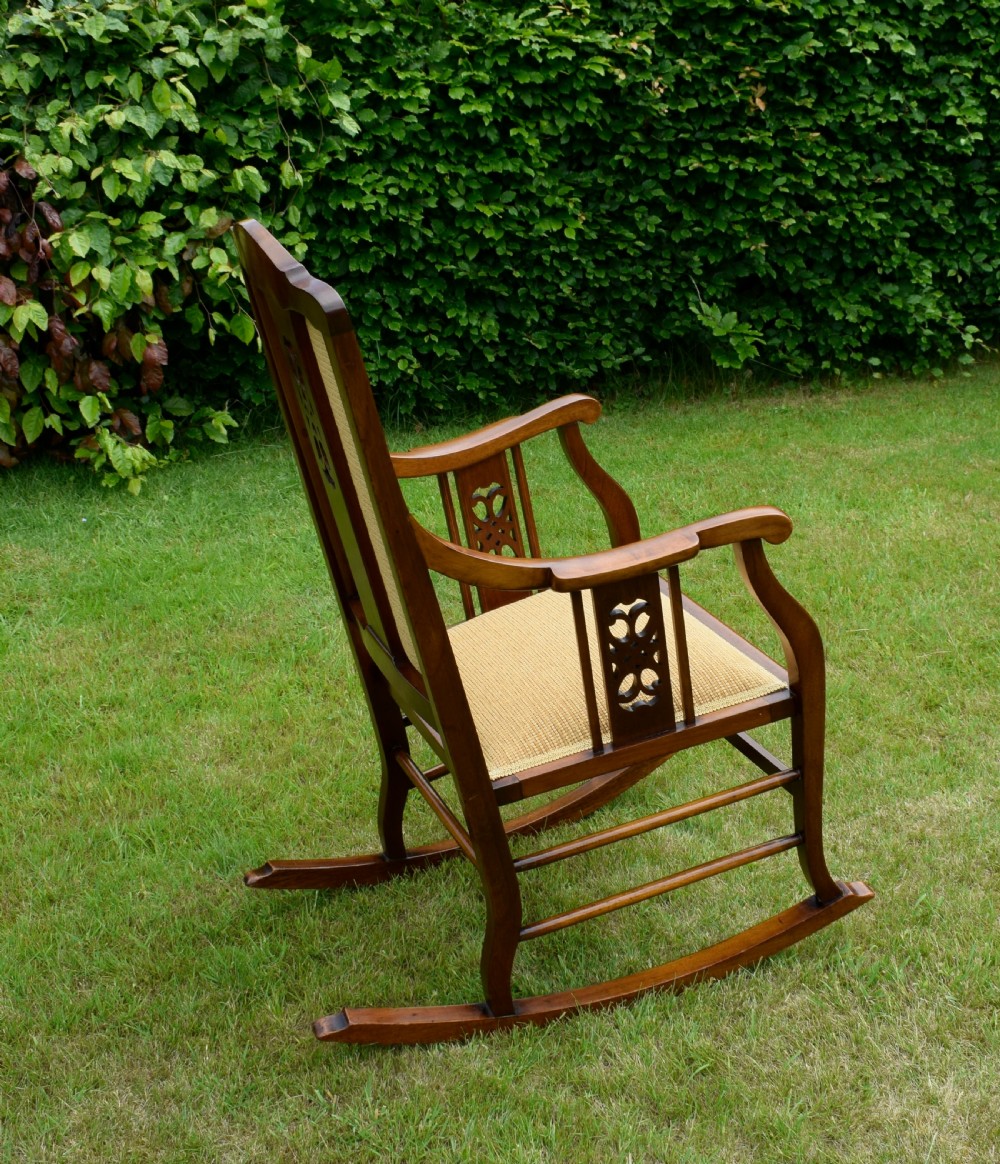 An Edwardian mahogany rocking chair featuring a crest rail with central marquetry inlay, double upholstered back panels, pierced fretwork splats, and light beige textured upholstery. View XXV
