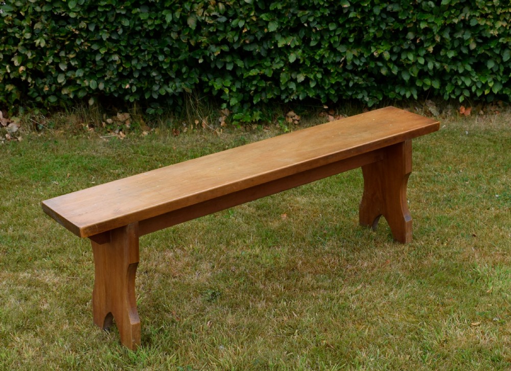 A long, simple wooden bench featuring a rectangular plank seat and cut-out trestle legs in a warm brown timber finish.