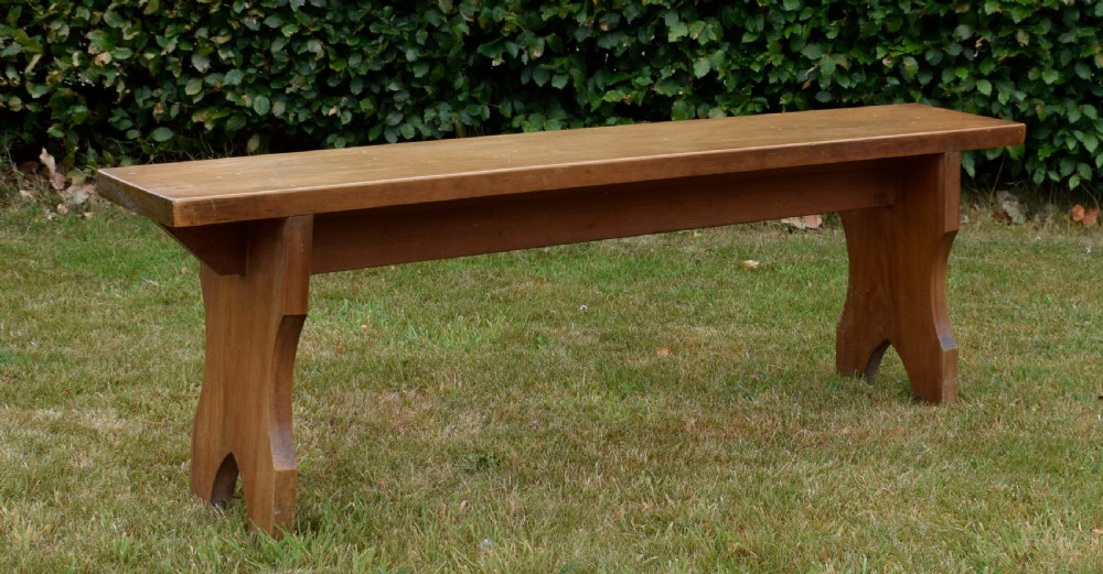 A long, simple wooden bench featuring a rectangular plank seat and cut-out trestle legs in a warm brown timber finish. View II