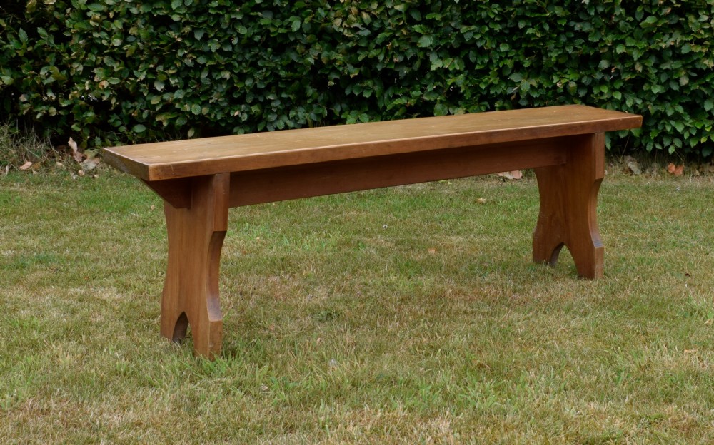 A long, simple wooden bench featuring a rectangular plank seat and cut-out trestle legs in a warm brown timber finish. View III