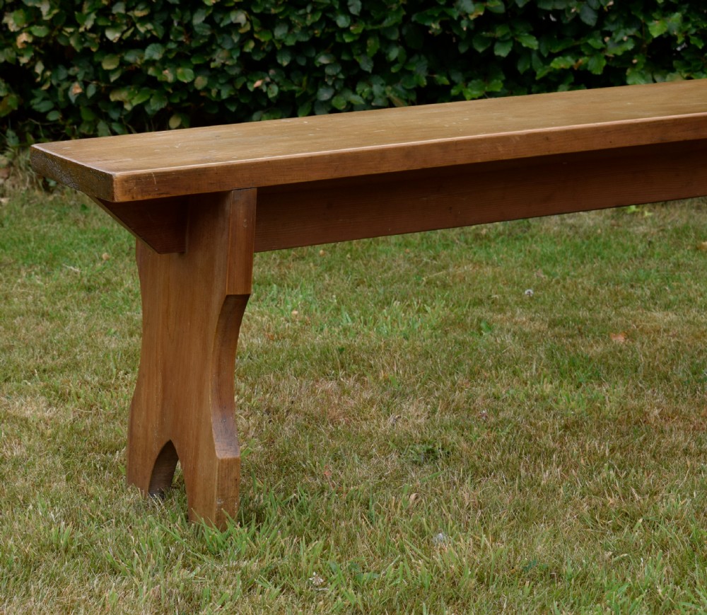 A long, simple wooden bench featuring a rectangular plank seat and cut-out trestle legs in a warm brown timber finish. View IV