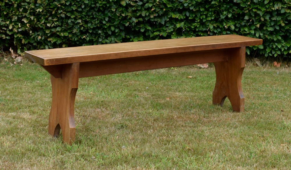 A long, simple wooden bench featuring a rectangular plank seat and cut-out trestle legs in a warm brown timber finish. View V