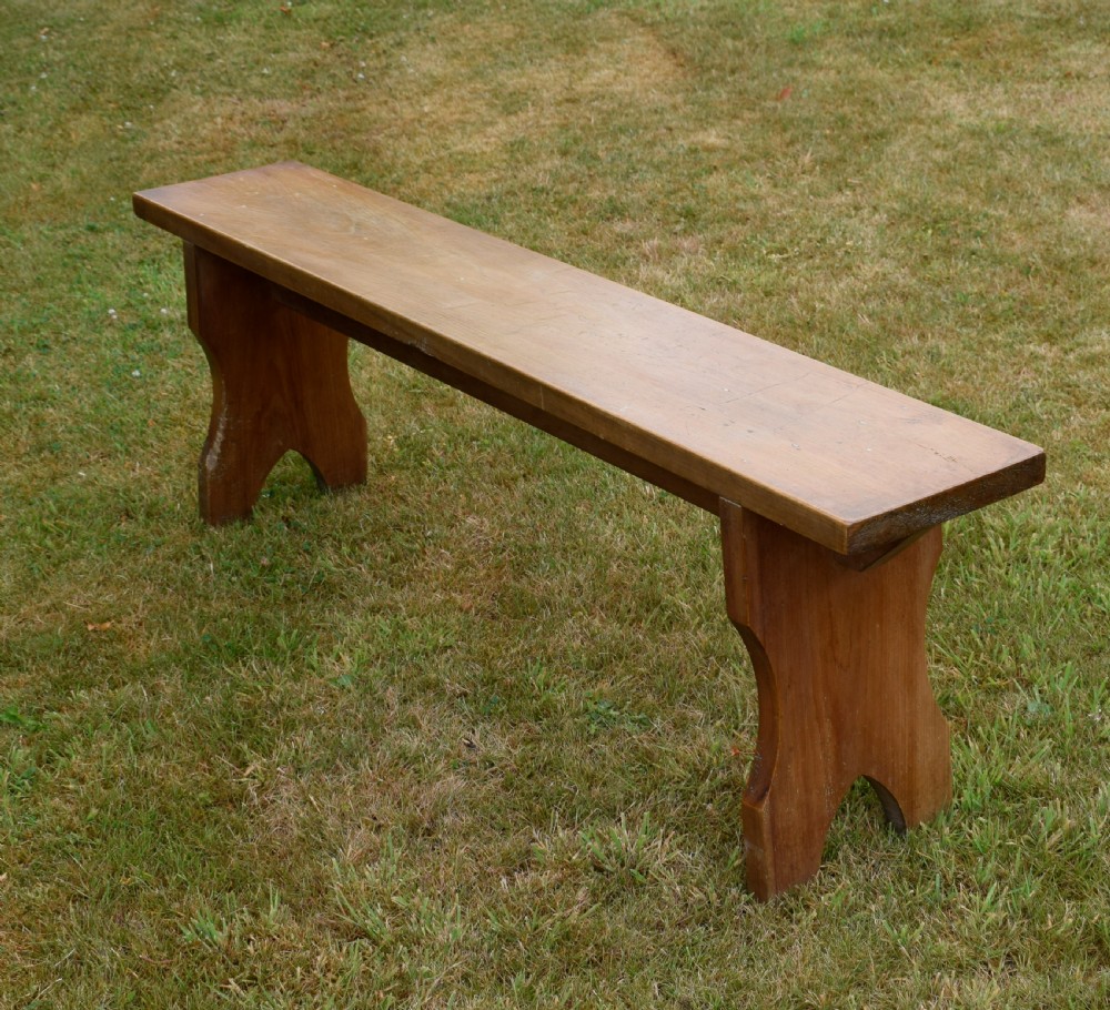 A long, simple wooden bench featuring a rectangular plank seat and cut-out trestle legs in a warm brown timber finish. View VI