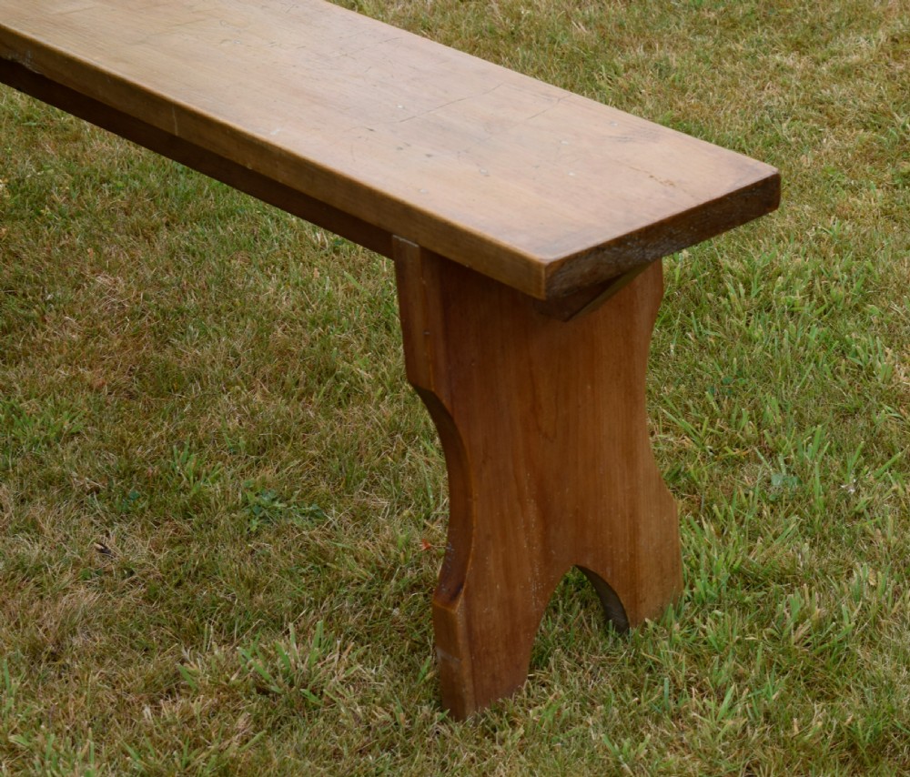 A long, simple wooden bench featuring a rectangular plank seat and cut-out trestle legs in a warm brown timber finish. View VII