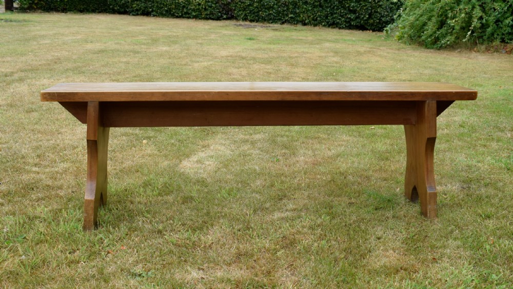 A long, simple wooden bench featuring a rectangular plank seat and cut-out trestle legs in a warm brown timber finish. View VIII