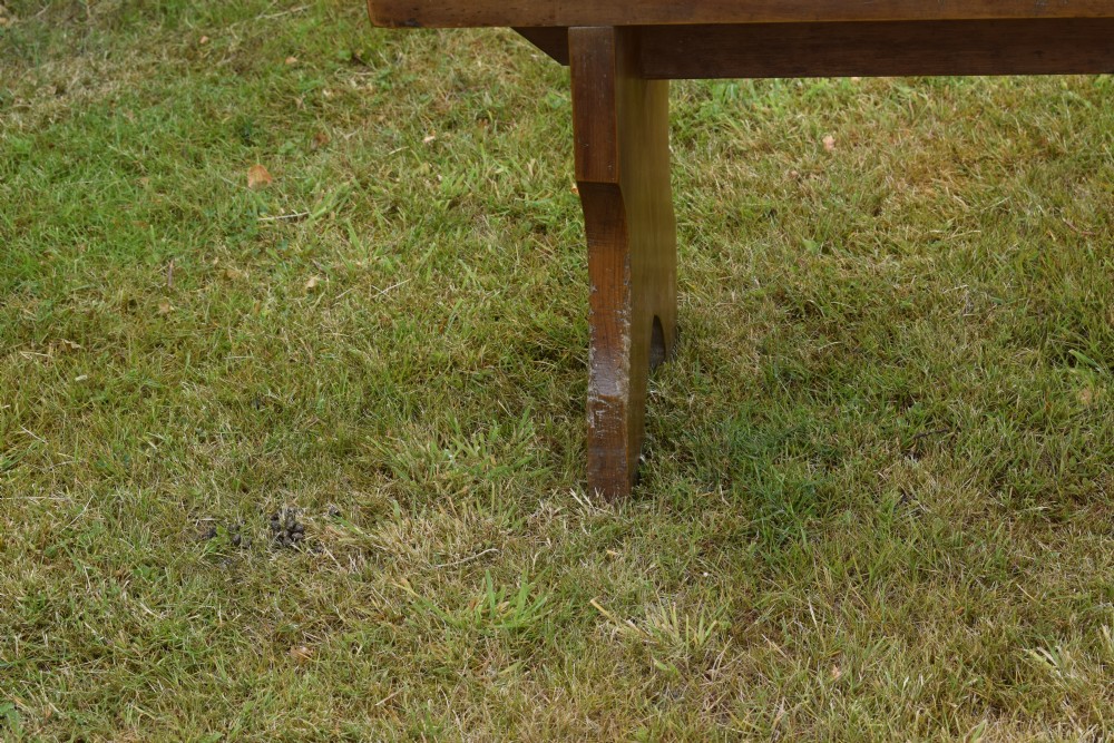 A long, simple wooden bench featuring a rectangular plank seat and cut-out trestle legs in a warm brown timber finish. View IX