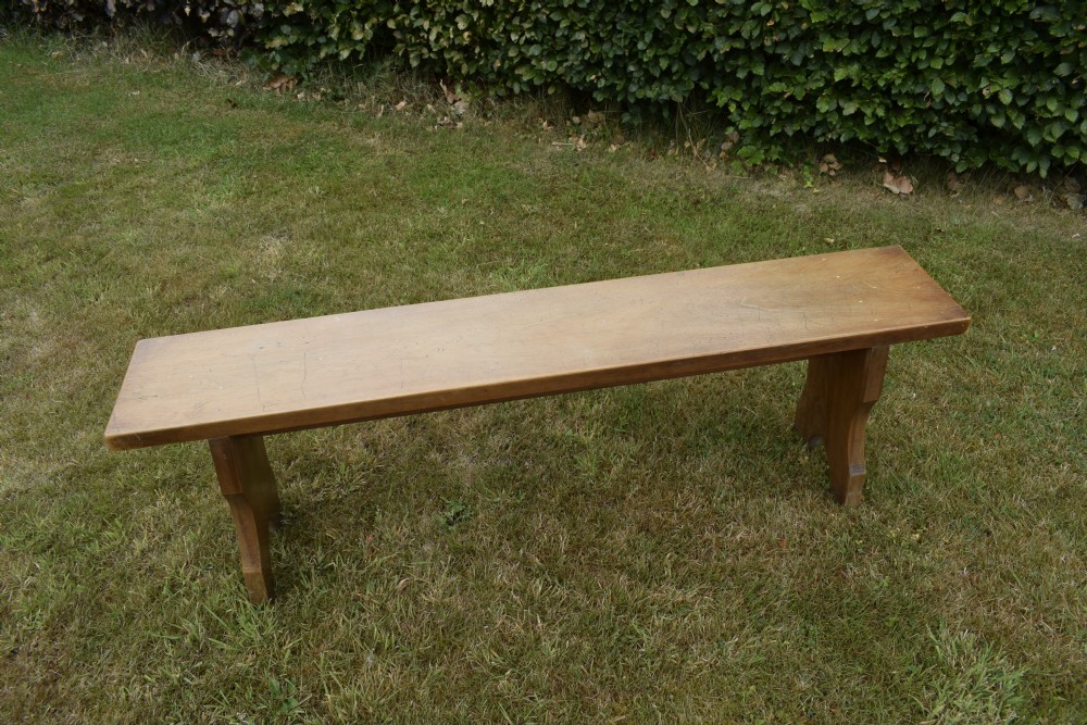 A long, simple wooden bench featuring a rectangular plank seat and cut-out trestle legs in a warm brown timber finish. View XII