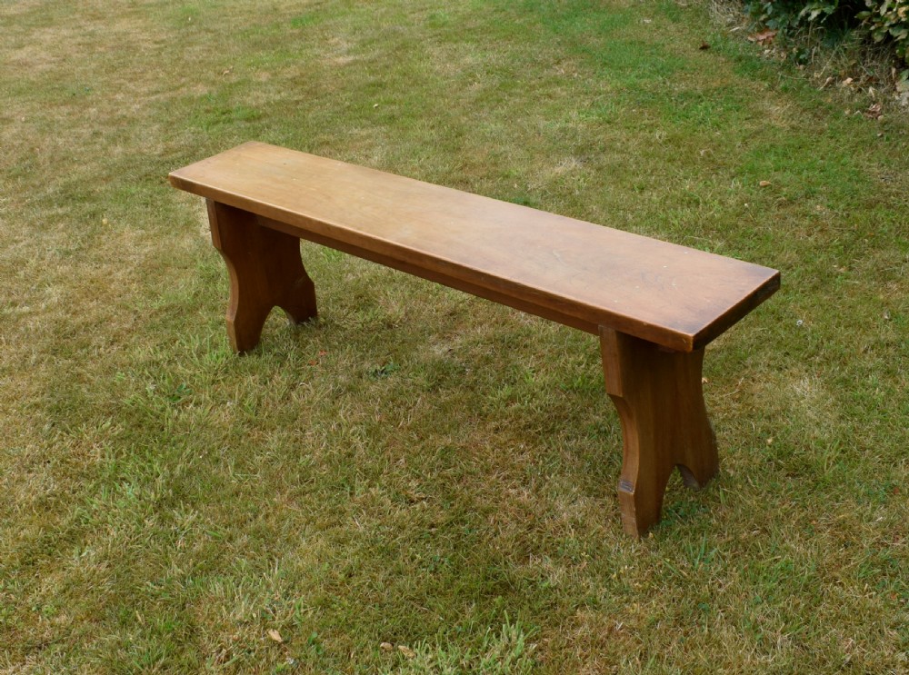 A long, simple wooden bench featuring a rectangular plank seat and cut-out trestle legs in a warm brown timber finish. View XIII