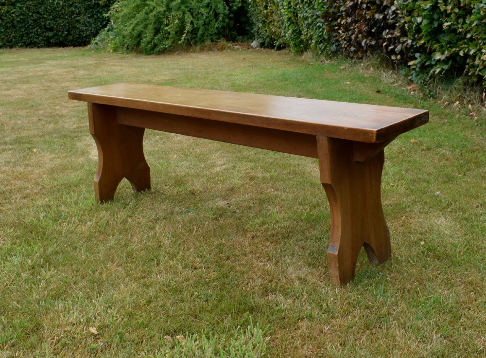 A long, simple wooden bench featuring a rectangular plank seat and cut-out trestle legs in a warm brown timber finish. View XIV