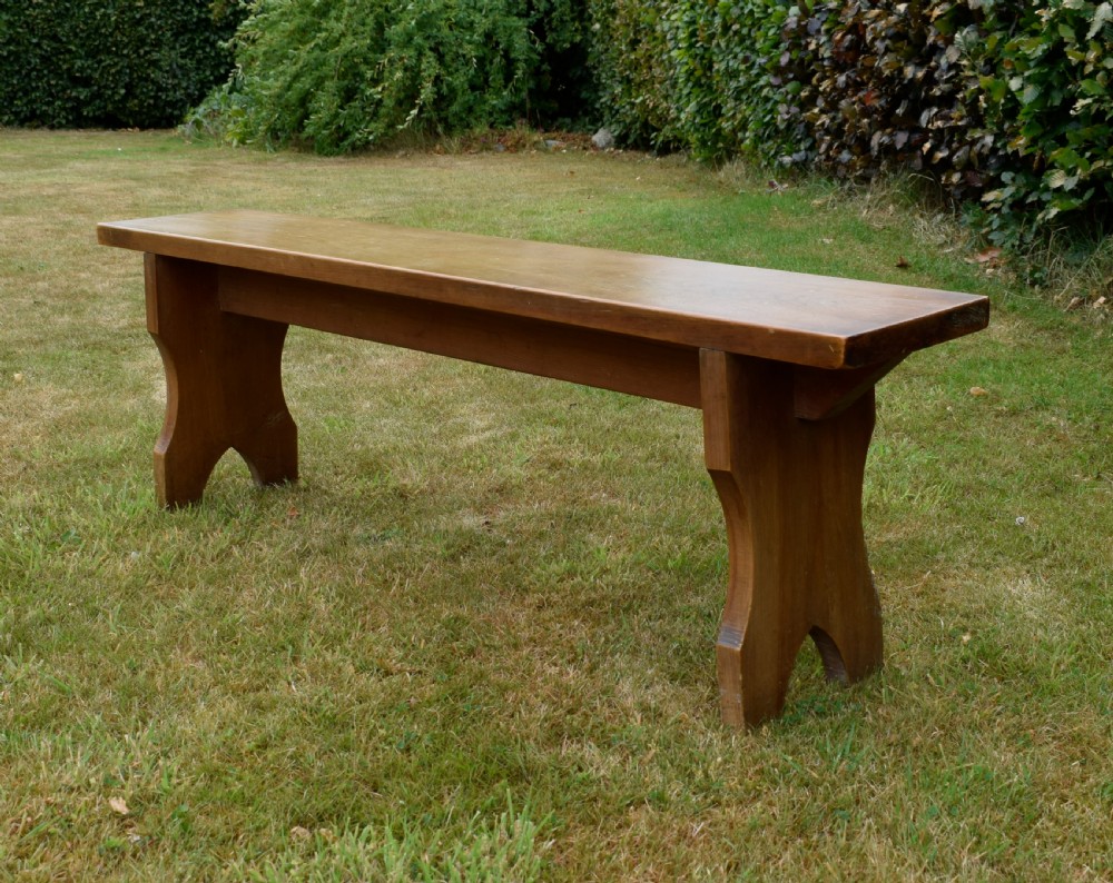 A long, simple wooden bench featuring a rectangular plank seat and cut-out trestle legs in a warm brown timber finish. View XV
