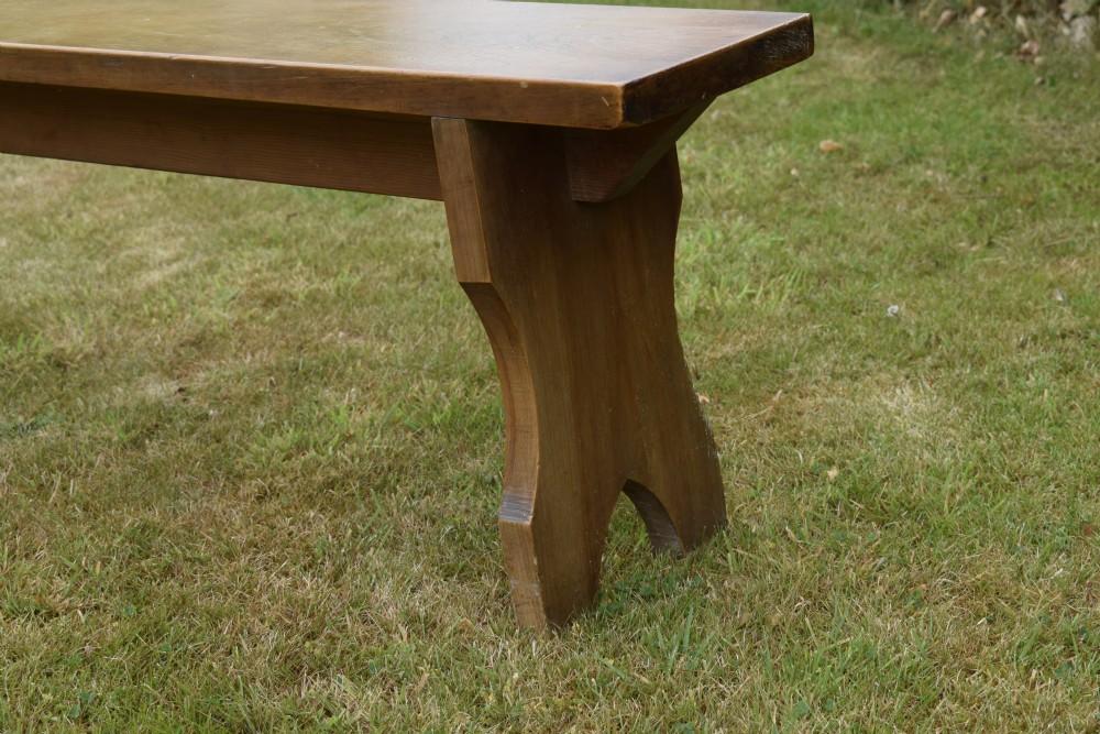 A long, simple wooden bench featuring a rectangular plank seat and cut-out trestle legs in a warm brown timber finish. View XVI