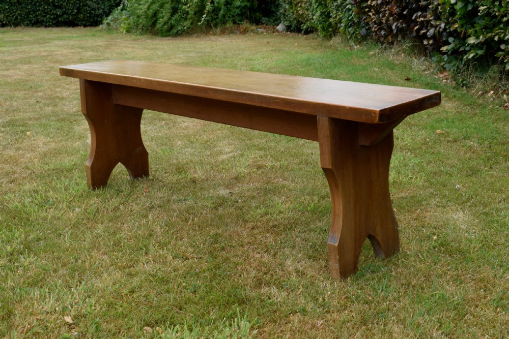 A long, simple wooden bench featuring a rectangular plank seat and cut-out trestle legs in a warm brown timber finish. View XVII