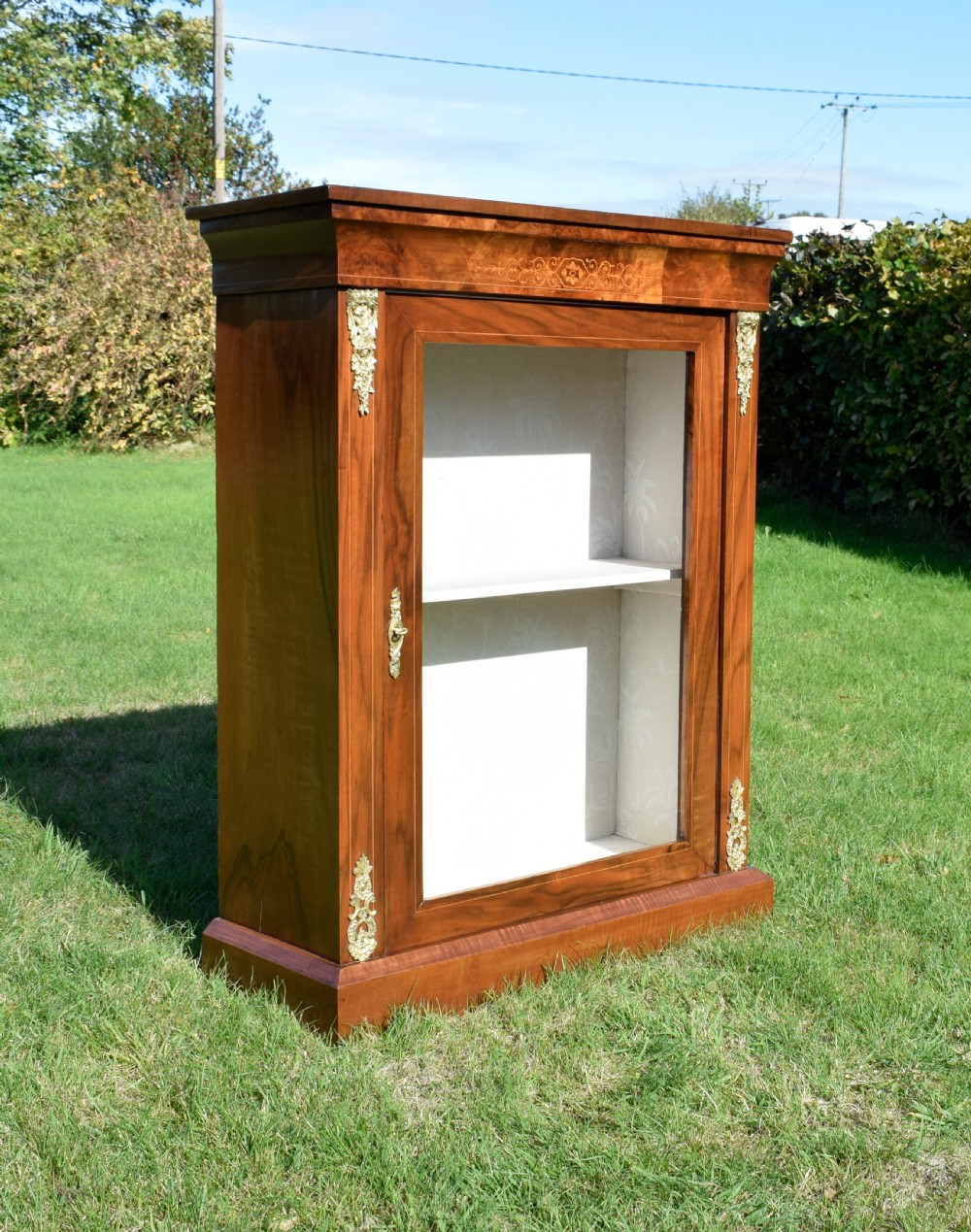 A Victorian warm brown walnut display cabinet featuring a single glazed door, floral wood inlay on the top frieze, and decorative brass mounts. View III