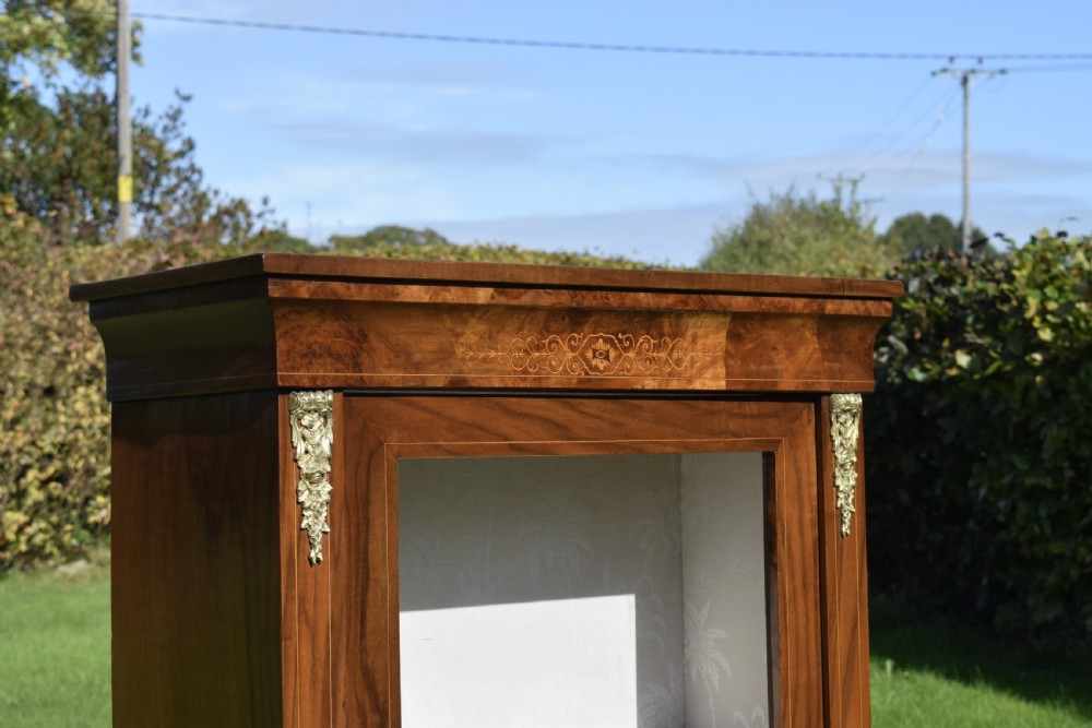 A Victorian warm brown walnut display cabinet featuring a single glazed door, floral wood inlay on the top frieze, and decorative brass mounts. View V