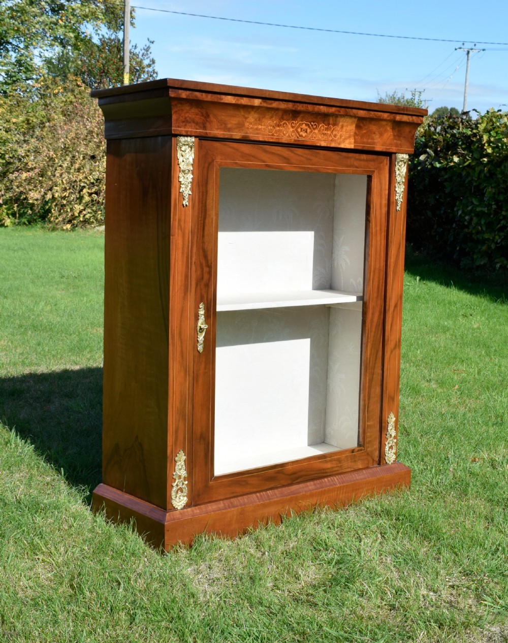 A Victorian warm brown walnut display cabinet featuring a single glazed door, floral wood inlay on the top frieze, and decorative brass mounts. View VI