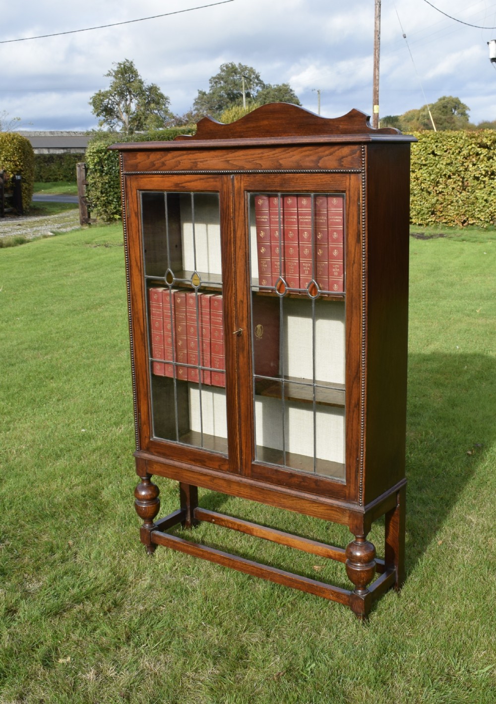 An early 20th-century oak bookcase featuring two leaded glass doors, a shaped top cresting rail, and turned legs joined by stretchers.
