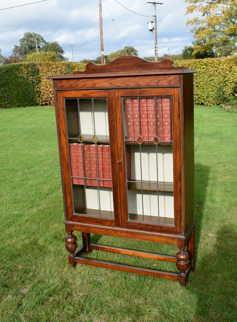 An early 20th-century oak bookcase featuring two leaded glass doors, a shaped top cresting rail, and turned legs joined by stretchers. View II