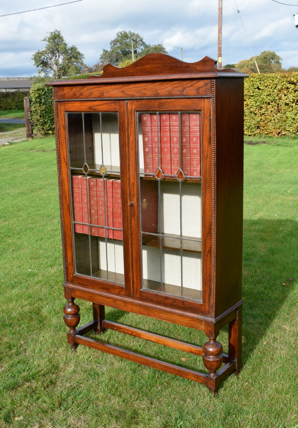 An early 20th-century oak bookcase featuring two leaded glass doors, a shaped top cresting rail, and turned legs joined by stretchers. View III