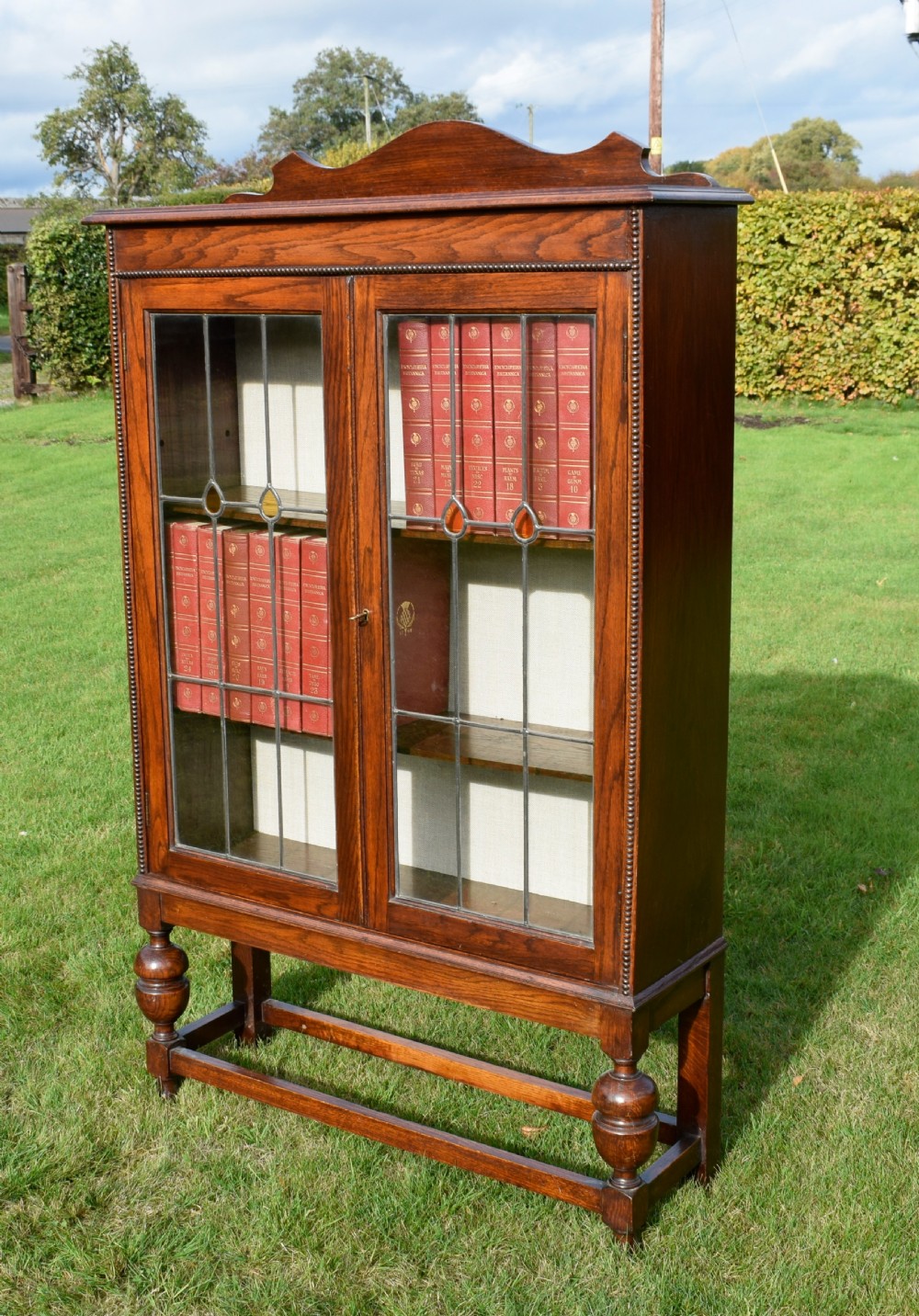 An early 20th-century oak bookcase featuring two leaded glass doors, a shaped top cresting rail, and turned legs joined by stretchers. View V