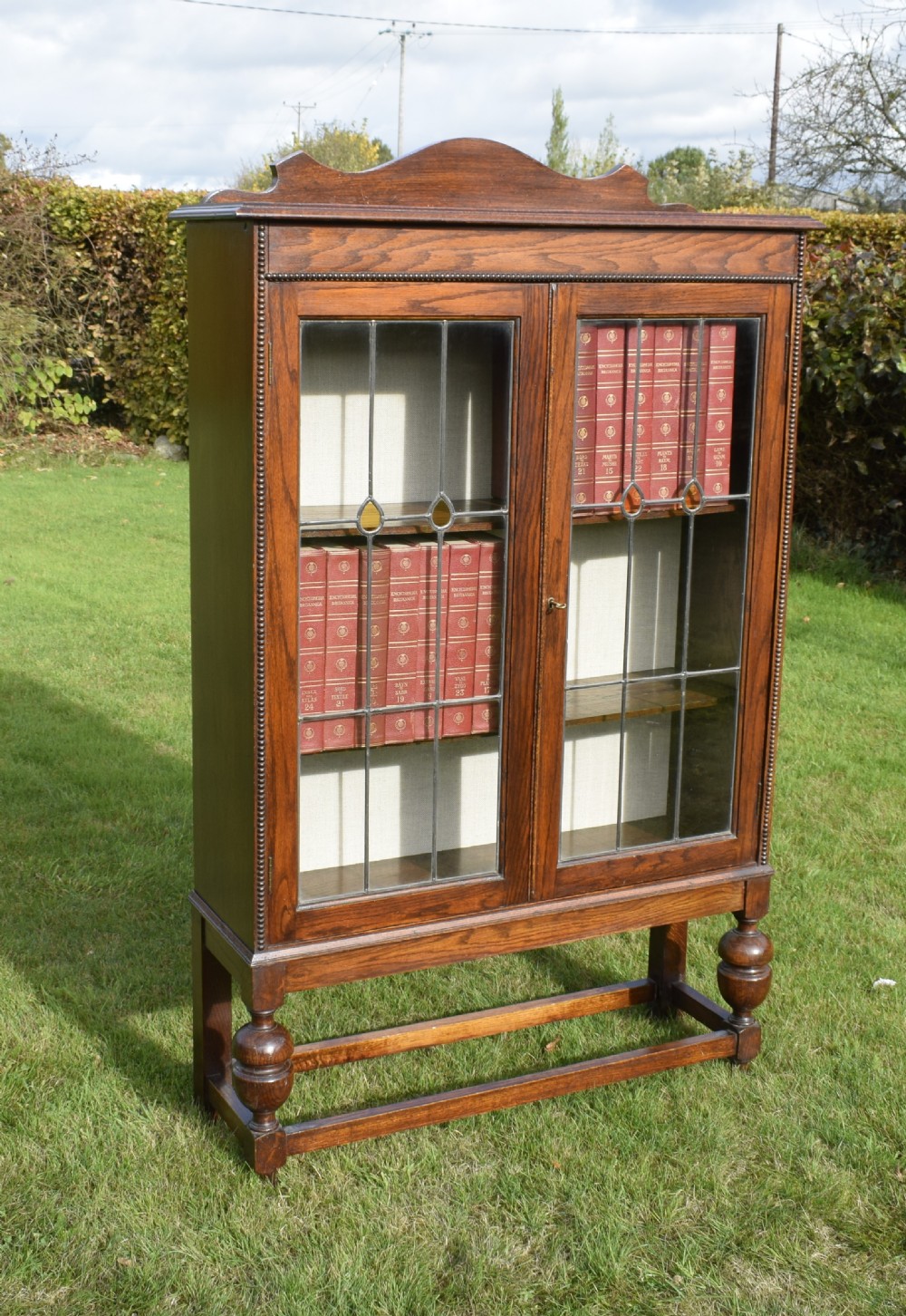An early 20th-century oak bookcase featuring two leaded glass doors, a shaped top cresting rail, and turned legs joined by stretchers. View VI