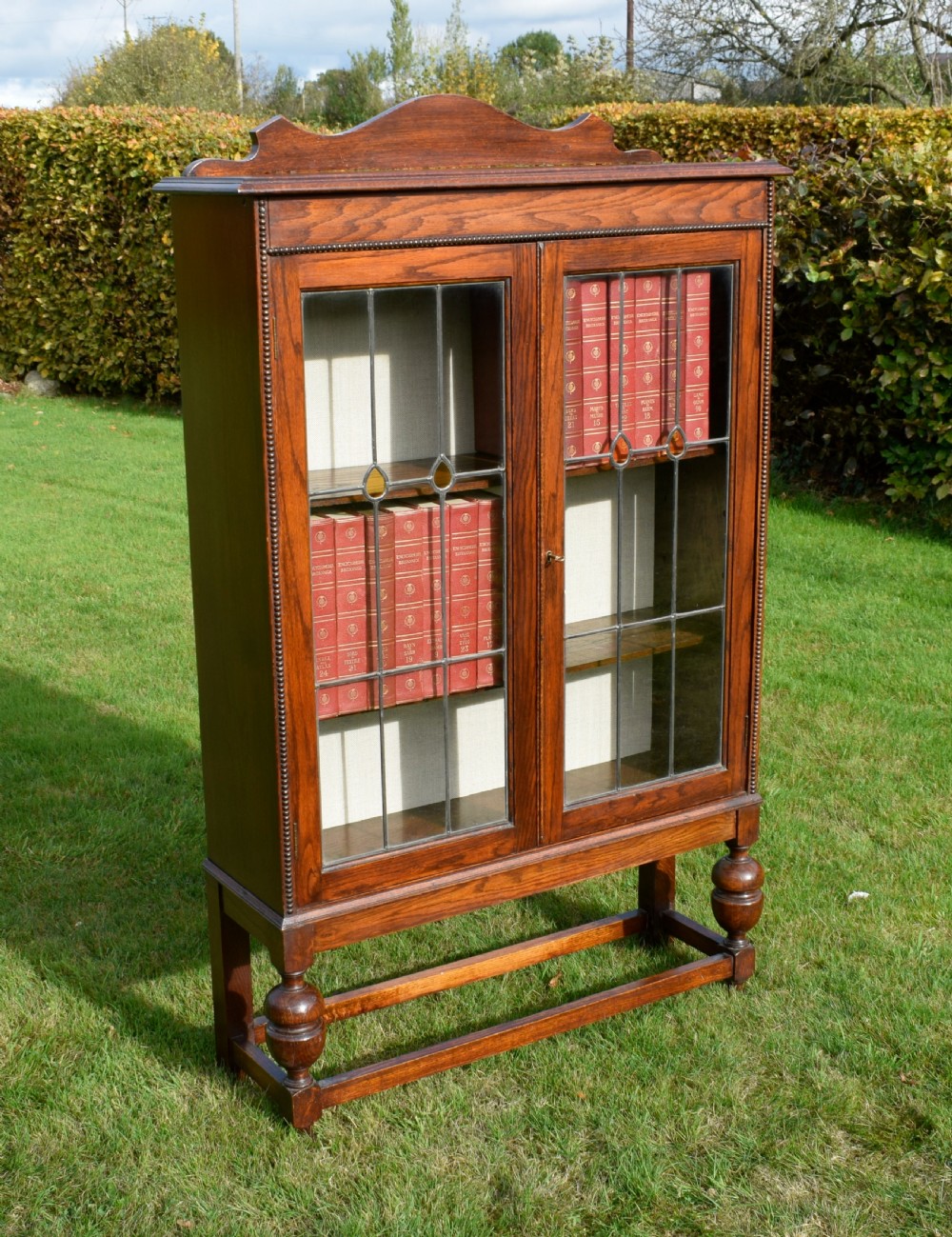 An early 20th-century oak bookcase featuring two leaded glass doors, a shaped top cresting rail, and turned legs joined by stretchers. View VII
