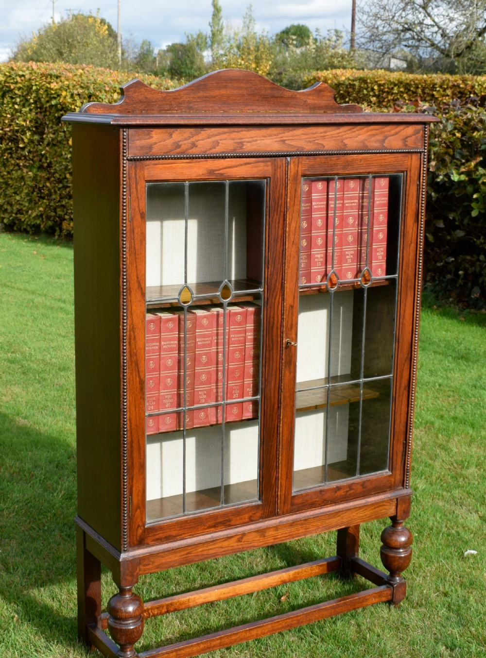 An early 20th-century oak bookcase featuring two leaded glass doors, a shaped top cresting rail, and turned legs joined by stretchers. View VIII