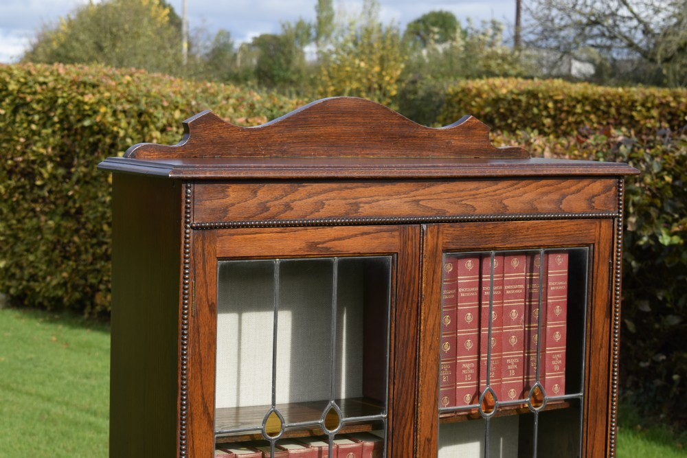 An early 20th-century oak bookcase featuring two leaded glass doors, a shaped top cresting rail, and turned legs joined by stretchers. View IX