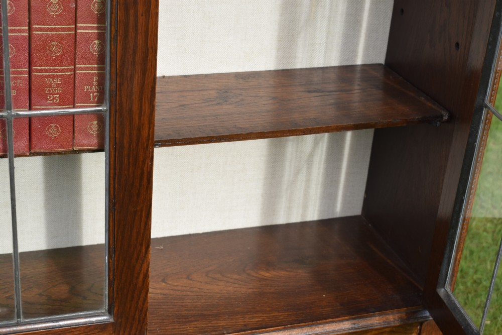 An early 20th-century oak bookcase featuring two leaded glass doors, a shaped top cresting rail, and turned legs joined by stretchers. View XIV