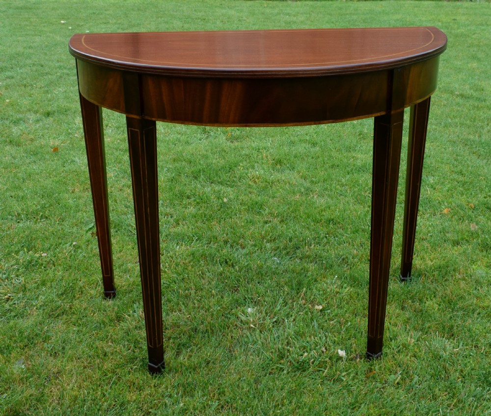 A mahogany demi-lune console table featuring light-coloured string inlay along the top perimeter, apron, and four square tapered legs.