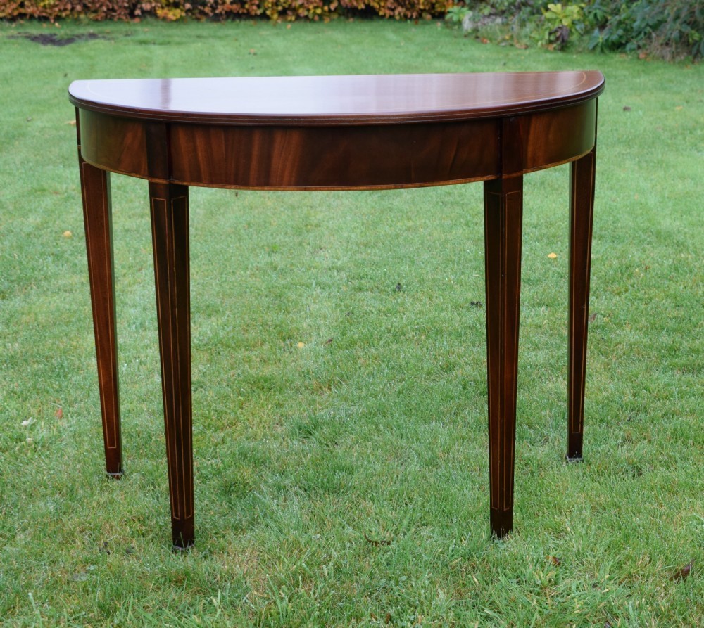A mahogany demi-lune console table featuring light-coloured string inlay along the top perimeter, apron, and four square tapered legs. View VI