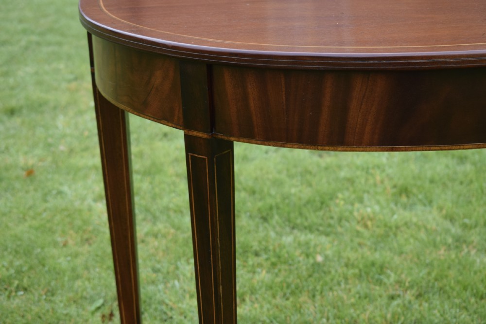 A mahogany demi-lune console table featuring light-coloured string inlay along the top perimeter, apron, and four square tapered legs. View X