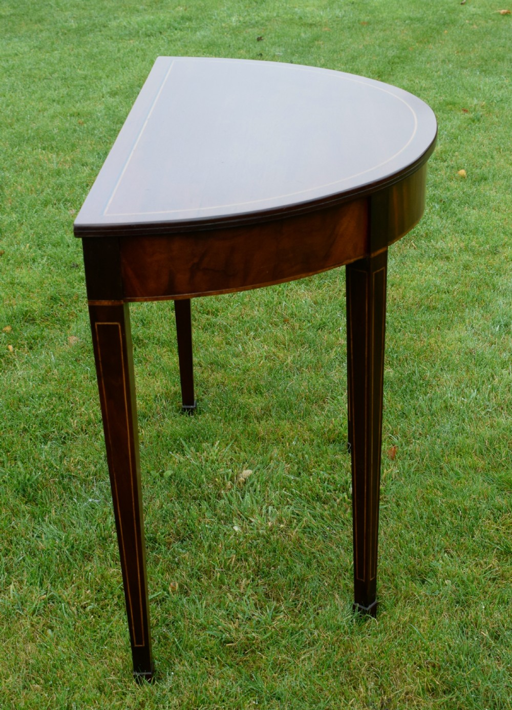 A mahogany demi-lune console table featuring light-coloured string inlay along the top perimeter, apron, and four square tapered legs. View XII