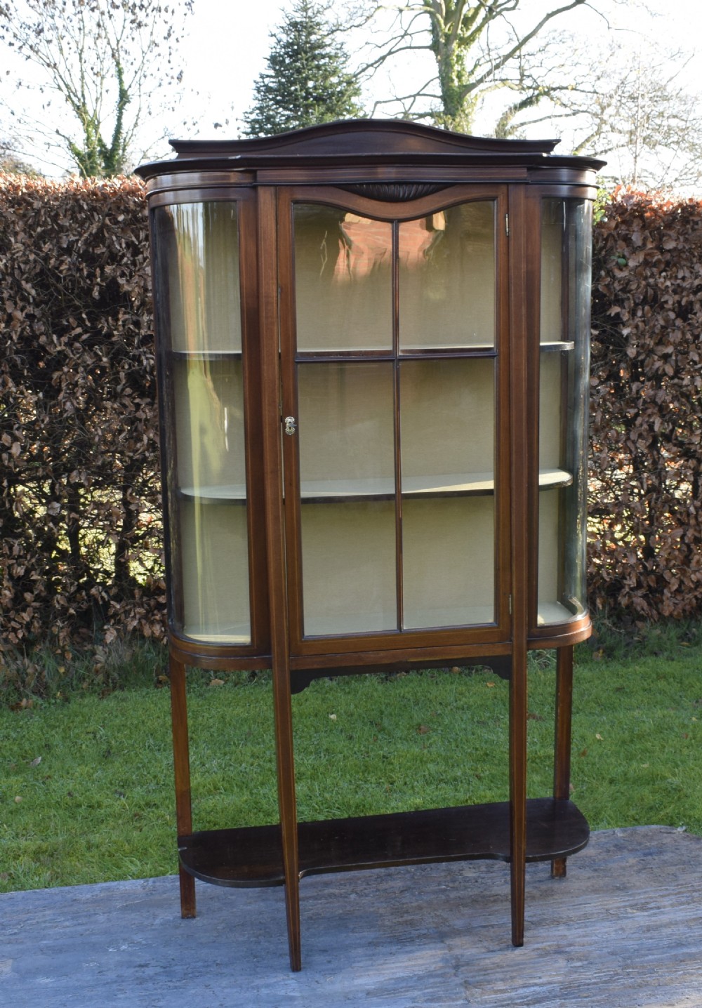 An Edwardian mahogany display cabinet featuring curved glass sides, line inlay details, a single glazed central door, and an undertier shelf supported by slender square legs. View VIII