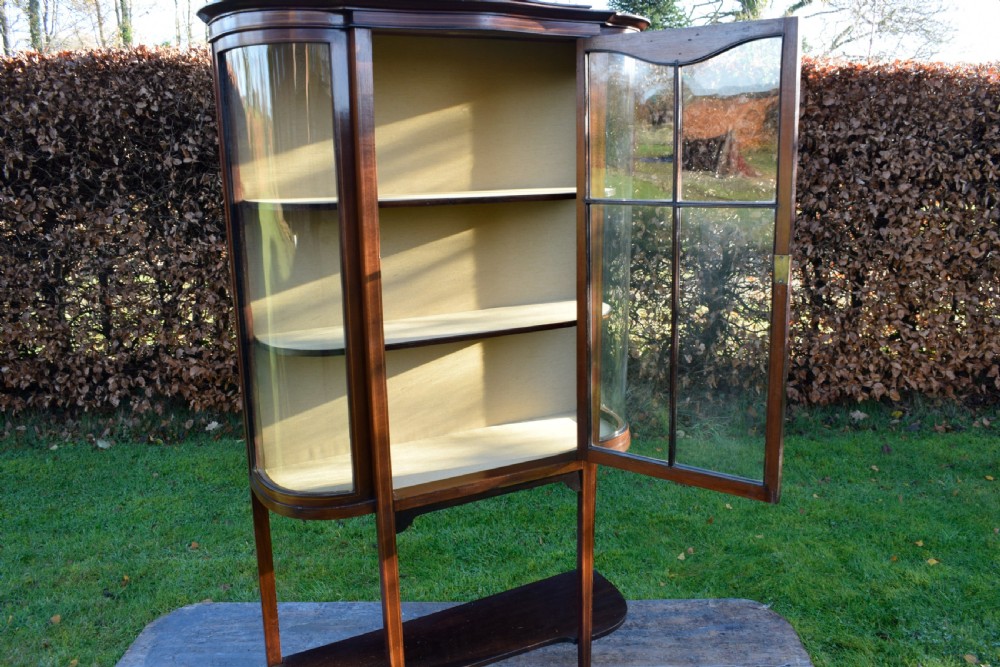 An Edwardian mahogany display cabinet featuring curved glass sides, line inlay details, a single glazed central door, and an undertier shelf supported by slender square legs. View IX