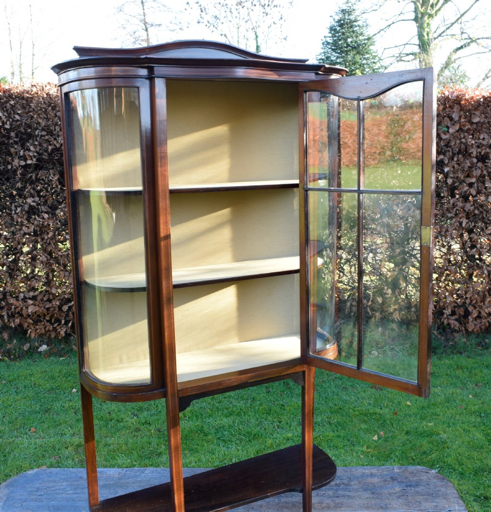 An Edwardian mahogany display cabinet featuring curved glass sides, line inlay details, a single glazed central door, and an undertier shelf supported by slender square legs. View X