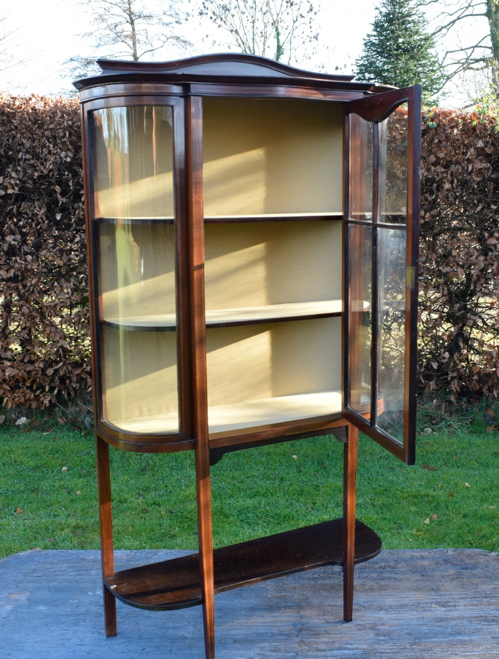An Edwardian mahogany display cabinet featuring curved glass sides, line inlay details, a single glazed central door, and an undertier shelf supported by slender square legs. View XI