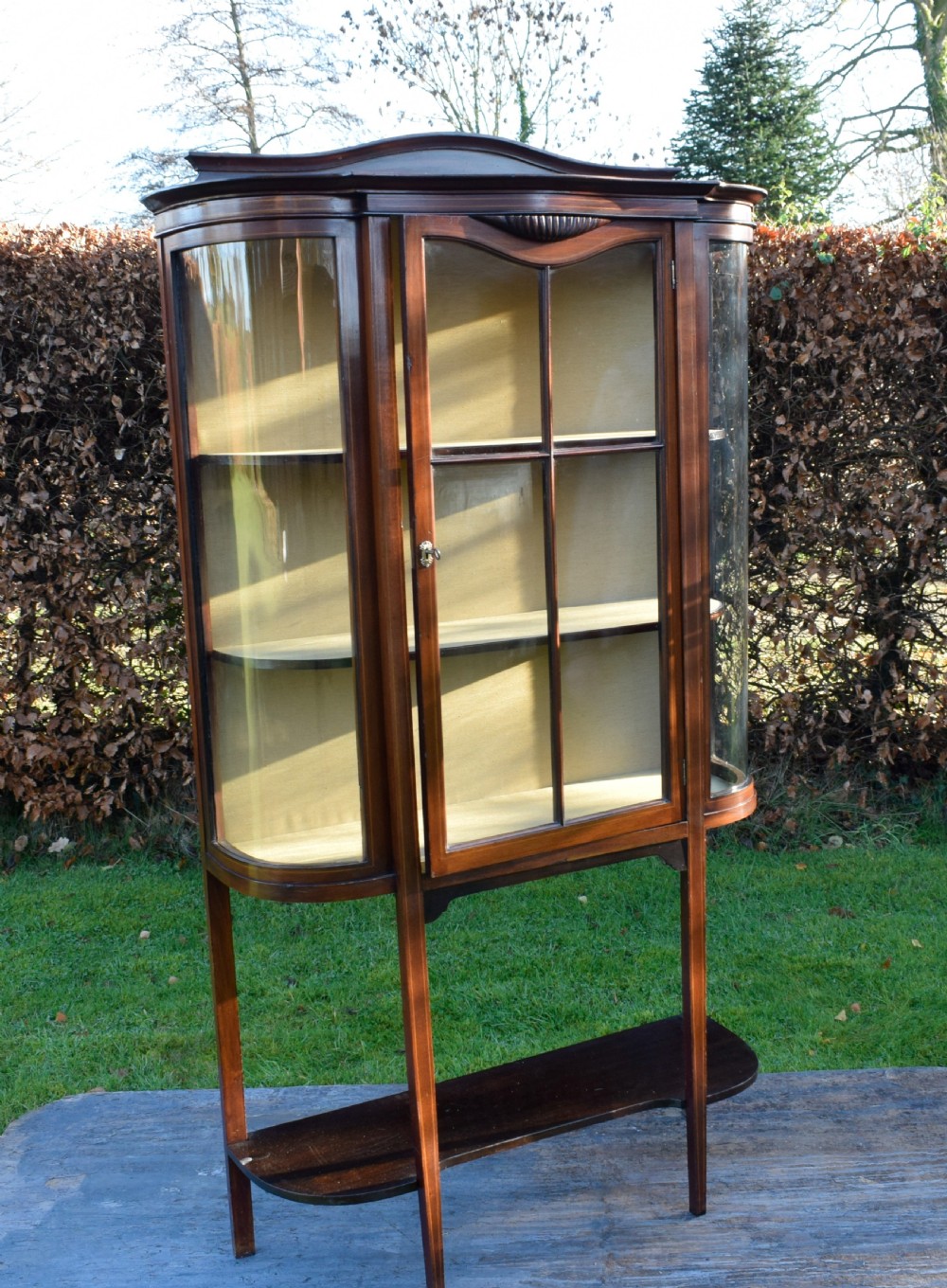 An Edwardian mahogany display cabinet featuring curved glass sides, line inlay details, a single glazed central door, and an undertier shelf supported by slender square legs. View XII