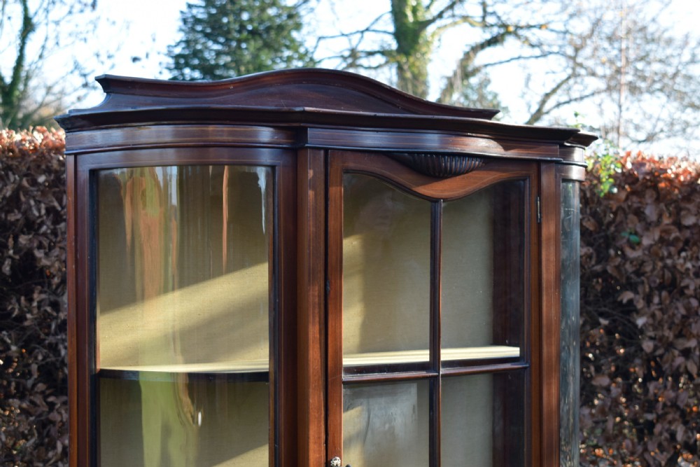 An Edwardian mahogany display cabinet featuring curved glass sides, line inlay details, a single glazed central door, and an undertier shelf supported by slender square legs. View XIV