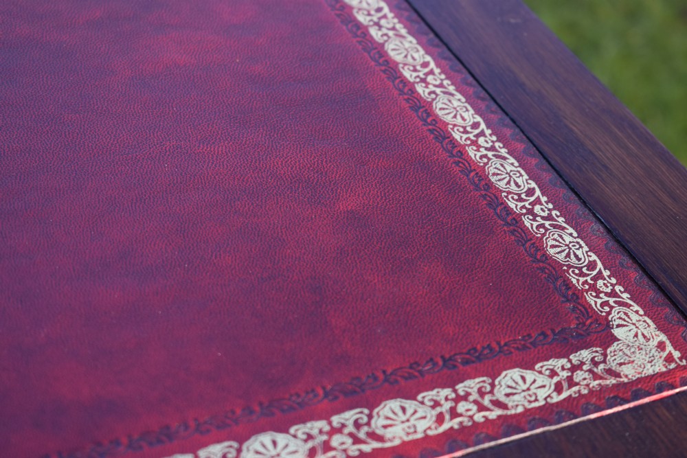 A rectangular Arts and Crafts oak writing table featuring a burgundy leather inset top with gold tooling and a lower H-stretcher. View X