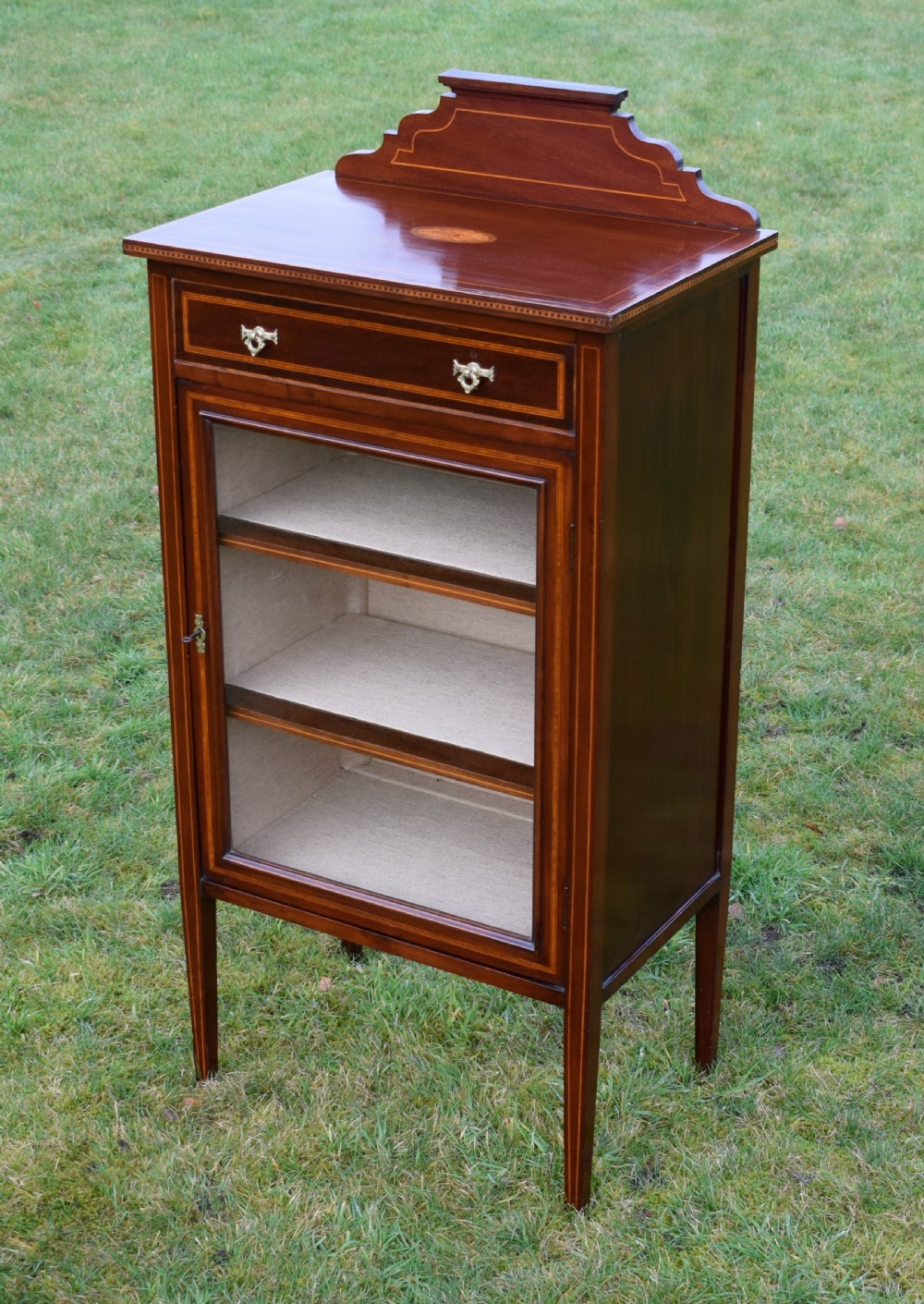 An Edwardian inlaid mahogany display cabinet featuring a shaped upstand gallery, a single top drawer with brass drop handles, and a glass-fronted door opening to fabric-lined interior shelves.
