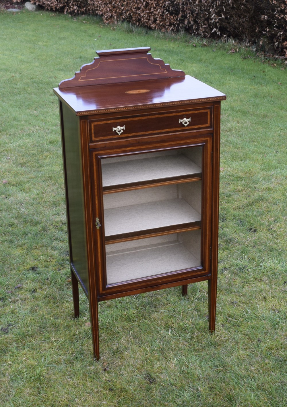 An Edwardian inlaid mahogany display cabinet featuring a shaped upstand gallery, a single top drawer with brass drop handles, and a glass-fronted door opening to fabric-lined interior shelves. View III