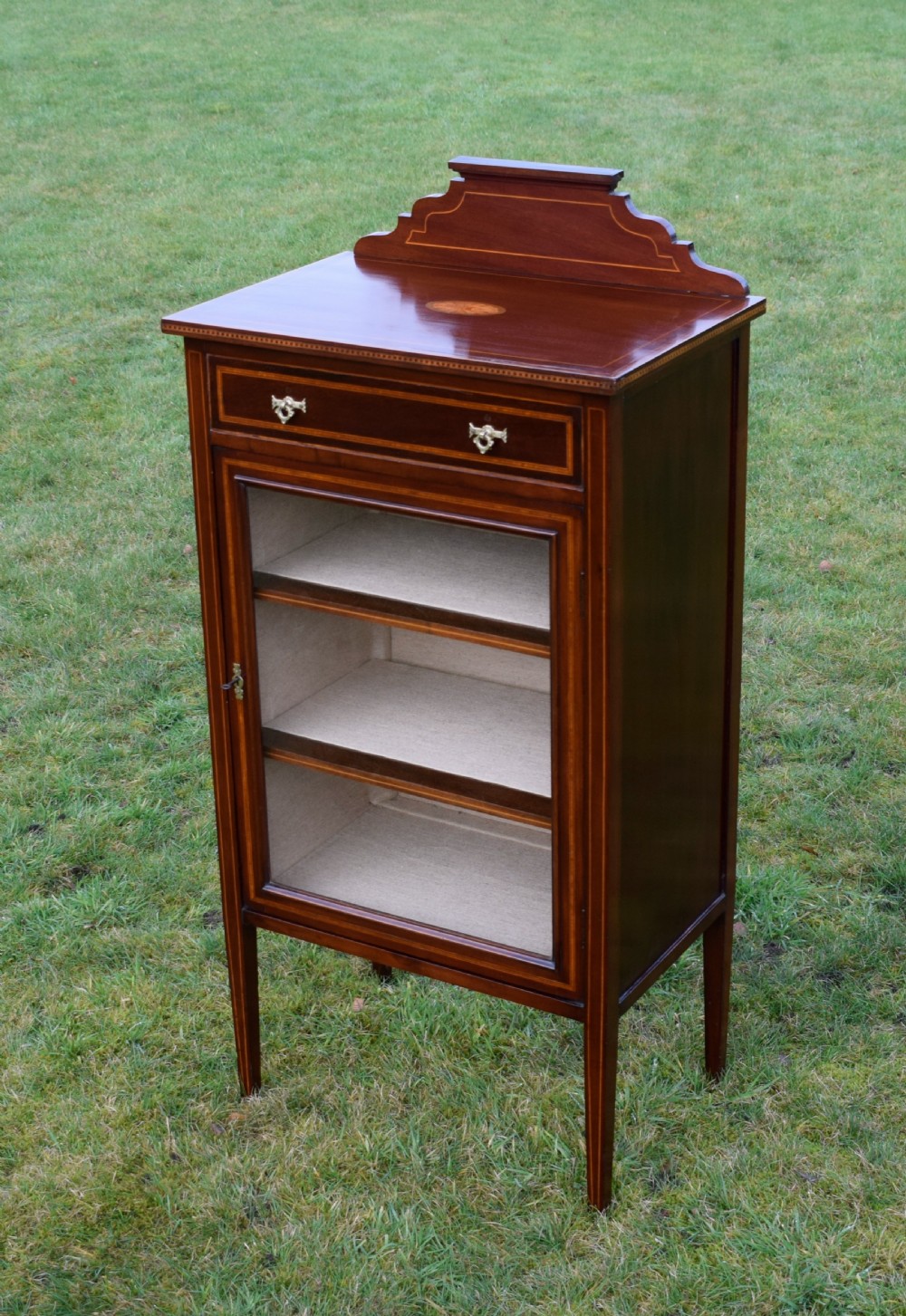 An Edwardian inlaid mahogany display cabinet featuring a shaped upstand gallery, a single top drawer with brass drop handles, and a glass-fronted door opening to fabric-lined interior shelves. View IV