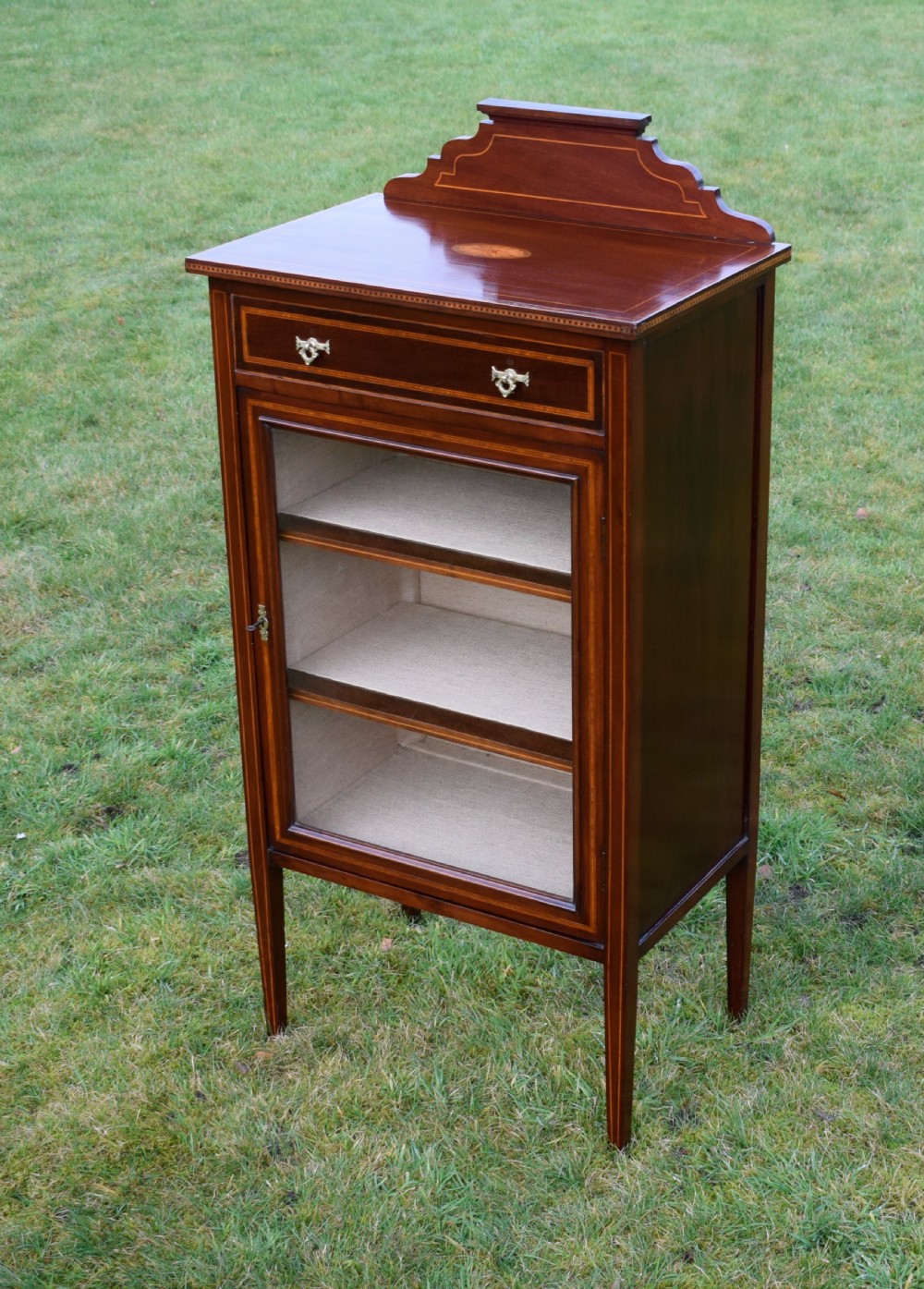 An Edwardian inlaid mahogany display cabinet featuring a shaped upstand gallery, a single top drawer with brass drop handles, and a glass-fronted door opening to fabric-lined interior shelves. View V