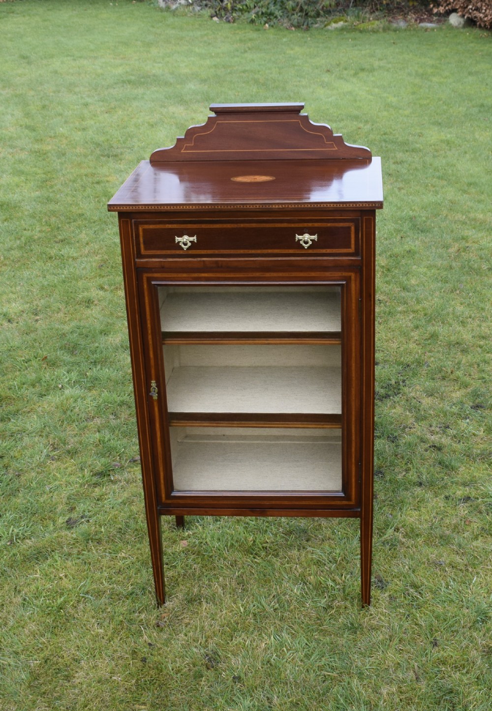 An Edwardian inlaid mahogany display cabinet featuring a shaped upstand gallery, a single top drawer with brass drop handles, and a glass-fronted door opening to fabric-lined interior shelves. View XI