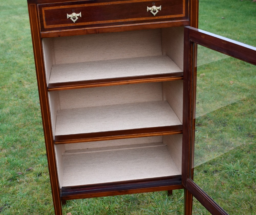 An Edwardian inlaid mahogany display cabinet featuring a shaped upstand gallery, a single top drawer with brass drop handles, and a glass-fronted door opening to fabric-lined interior shelves. View XIII