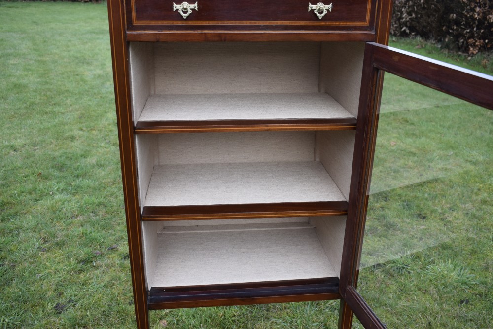 An Edwardian inlaid mahogany display cabinet featuring a shaped upstand gallery, a single top drawer with brass drop handles, and a glass-fronted door opening to fabric-lined interior shelves. View XVI