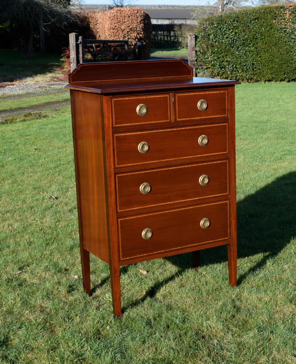 An Edwardian mahogany tall chest of drawers featuring fine string inlay, brass ring handles, five drawers, a low raised backboard, and square tapered legs.