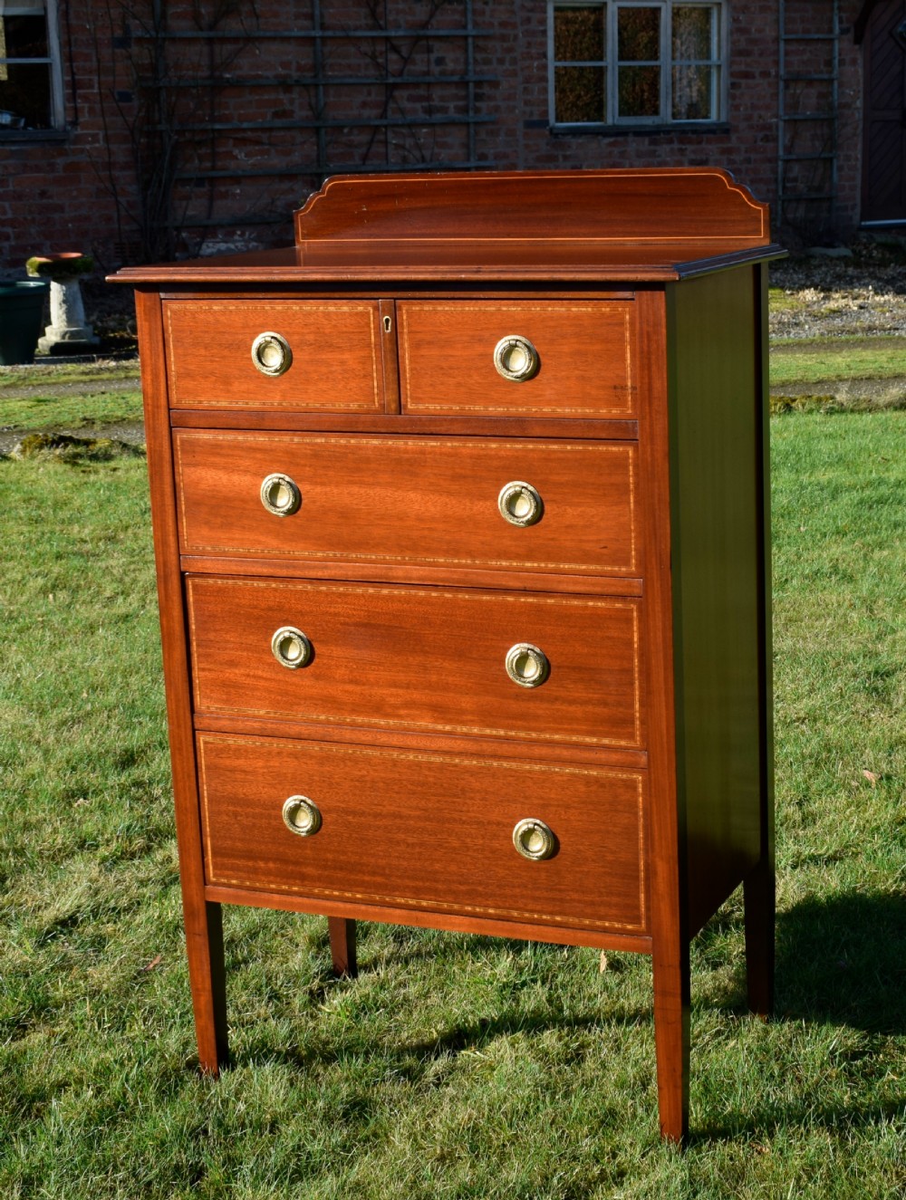 An Edwardian mahogany tall chest of drawers featuring fine string inlay, brass ring handles, five drawers, a low raised backboard, and square tapered legs. View V