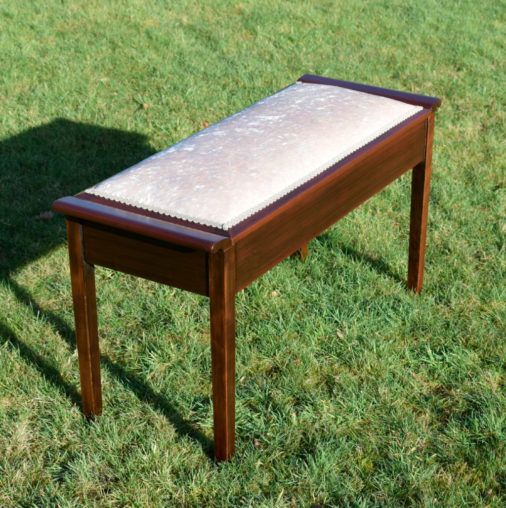 An Art Deco dark mahogany duet piano stool featuring a cushioned pale pink velvet seat with braided gimp trim and straight tapered legs.