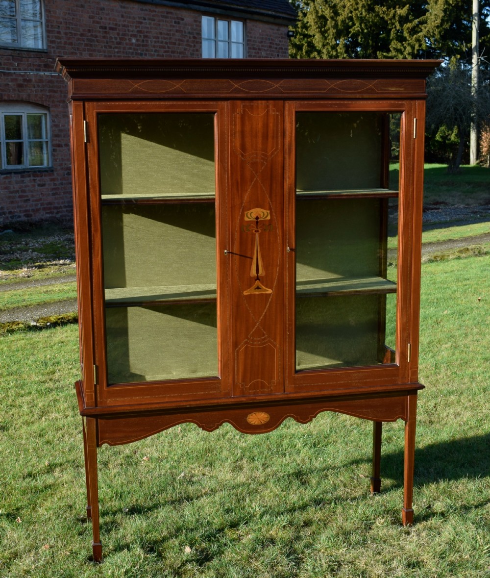 An early 20th-century mahogany Art Nouveau display cabinet featuring glazed doors, central inlaid botanical marquetry, and a fabric-lined interior on slender legs.